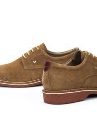 Chaussures oxford en daim marron avec un nez arrondi, des lacets châtaigne clair et un accent de talon bordeaux. La semelle extérieure présente un motif de crampons contrastant.