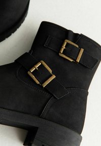 Czarne botki za kostkę z grubą podeszwą i dwoma mosiężnymi klamrami na jasnym tle.