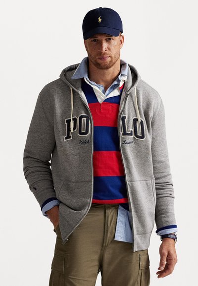 Grijze rits hoodie met "POLO" opdruk, gedragen over een rood en blauw gestreept polo-t-shirt. Een blauwe pet en kaki cargobroek maken de look compleet.