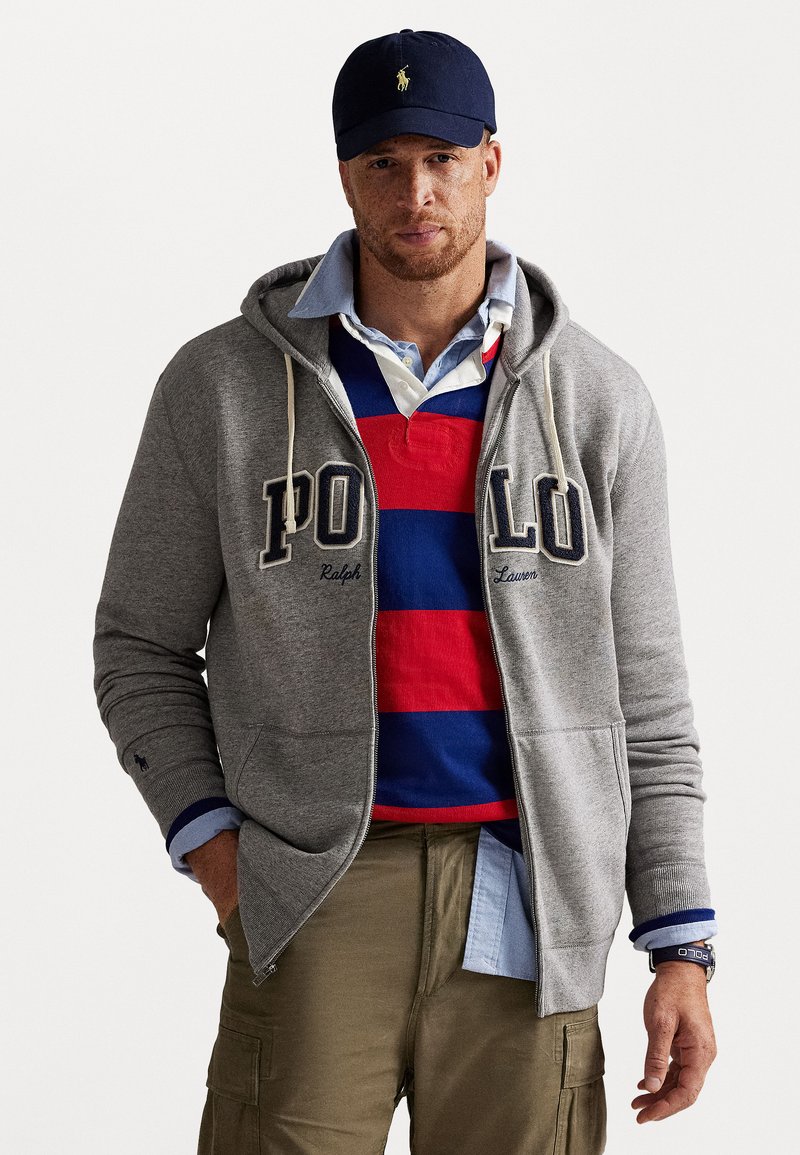 Sweatshirt gri cu fermoar, cu inscripția "POLO", purtat peste un tricou polo cu dungi roșii și albastre. O șapcă albastră și pantaloni cargo kaki completează ținuta.