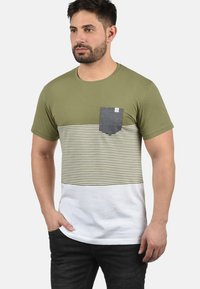 Camiseta de manga corta en verde oliva con una sección central a rayas horizontales en beige y oliva, con un bolsillo en el pecho de color gris oscuro y un borde blanco.