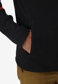 Mano parzialmente dentro della tasca laterale zip della giacca nera a maniche lunghe indossata con pantaloni marroni.