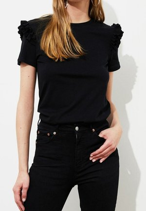 Kobieta ubrana w czarny krótki top z falbanami na ramionach oraz czarne, wysokie spodnie dżinsowe, ręka oparta na biodrze.