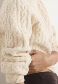 Personne portant un pull crème à torsades tenant son ventre, suggérant un malaise ou une douleur.