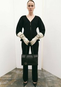 Čierny priliehavý top s výstrihom do V a dlhými rukávmi, v kombinácii s čiernymi nohavicami. Modelka nosí krémové rukavice a drží čiernu kabelku. Špicaté podpätky.
