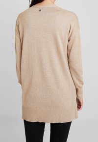 Beige långärmad tröja med rund halsringning, ribbad nederkant och sidenslitsar, tillverkad i mjukt stickat tyg. Liten logotyp vid bakre halsringningen.