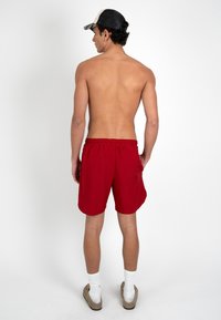 Jeune homme portant une casquette noire et blanche, un short rouge, des chaussettes blanches et des chaussures marron sans lacets, debout de dos.