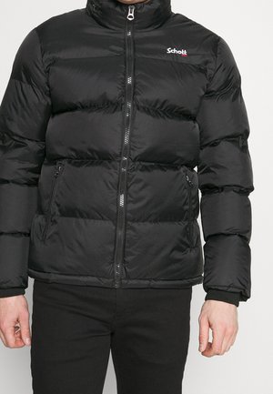 Man wearing black puffer jacket with front zipper and zipped side pockets, logo on left chest, paired with black pants, against plain background.