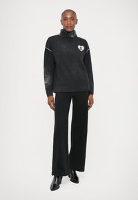 Suéter preto com gola alta, logotipo de coração e textura suave. Combinado com calças largas pretas e botas de bico fino.