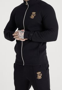 Tuta nera con giacca con zip e pantaloni abbinati. Presenta un logo dorato sul petto e sulla coscia. Tessuto elastico con texture liscia.