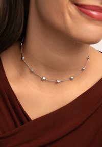 Collar de plata que presenta diseños alternos de corazones y cuentas redondas en una fina cadena, sobre un fondo de tela marrón oscuro.