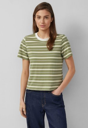 Woman wearing olive green and white striped short-sleeve shirt and dark blue jeans, standing with one hand in pocket against plain background.