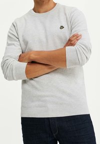 Ljusgrå tröja med strukturerat mönster, ribbade muddar och fåll. Liten gul broderad detalj på bröstet. Bärs med mörka jeans.