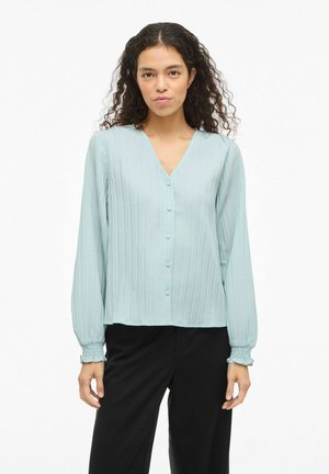 Hele sinine volditud pluus, millel on V-kaelus ja nööpidega kinnis. Pikad, keha järgivad varrukad, millel on elastsed varrukaservad. Sile tekstuur ja lõdvestunud lõige.