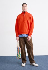 Pulôver de fleece laranja com um colarinho alto e meio zíper, sobre uma camisa azul-clara. Calças castanhas e ténis brancos. Segurando uma tote com padrão.