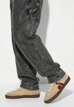 Persona con vaqueros negros desteñidos y zapatillas beige con textura de lana y suela marrón, sobre un fondo claro y liso.