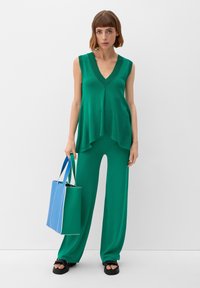 Groene mouwloze top met een V-hals, gecombineerd met brede groene broeken; houdt een twee-kleurige tas in blauw en groen met witte accenten vast.