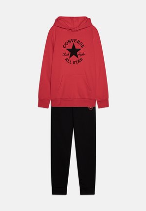 Roter Hoodie mit einem schwarzen "Converse All Star"-Logo und Stern-Design; kombiniert mit schwarzen Jogginghosen, die ein kleines Logo am Bein haben.