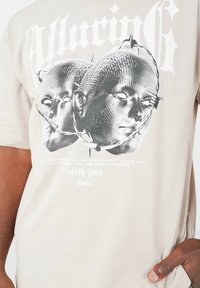 Beige bomulds t-shirt med et grafisk design af to stiliserede hoveder. Teksten nedenunder lyder: "Hvad du frygter mest... sjette juni, Paris."