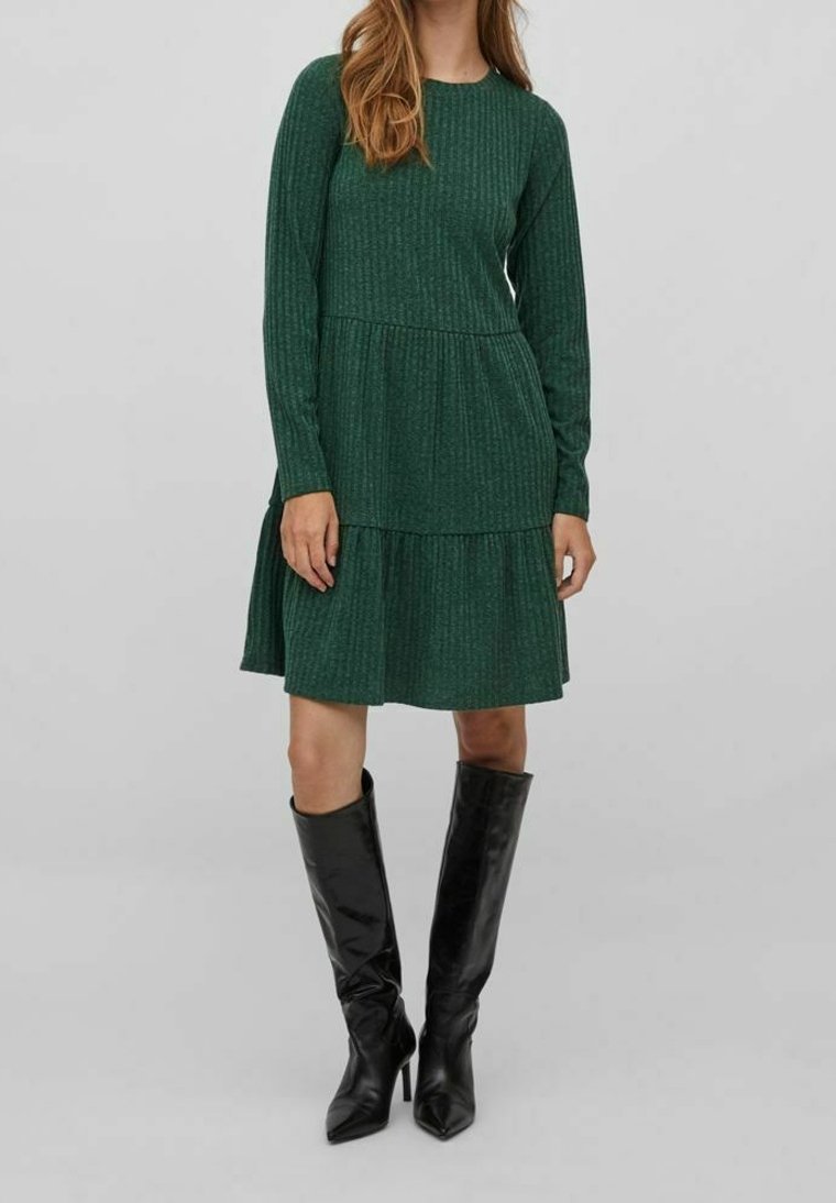 Femme portant une robe côtelée verte foncée à manches longues et des bottes noires montantes à talons, debout devant un fond gris uni.