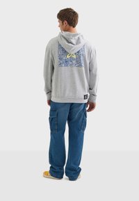 Sudadera con capucha gris y diseño gráfico azul en la parte trasera, combinada con pantalones cargo azul. Presenta un corte relajado y puños acanalados.