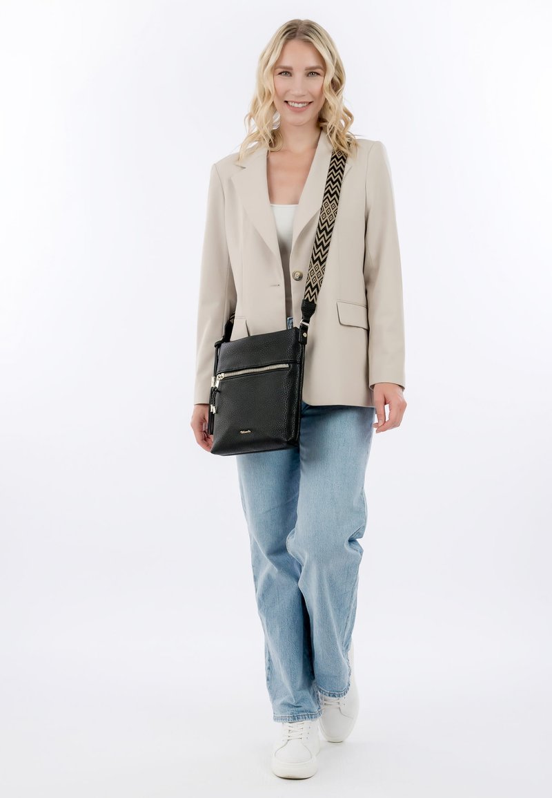 Glunderende vrouw die loopt, gekleed in een beige blazer, wit shirt, lichtblauwe spijkerbroek, witte sneakers, en een zwart crossbody tasje met een patroon op de band.