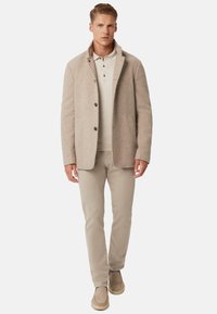 Hombre vestido con un abrigo de lana beige sobre una camisa polo beige clara, con pantalones beige y zapatos slip-on marrón claro, de pie mirando hacia adelante.