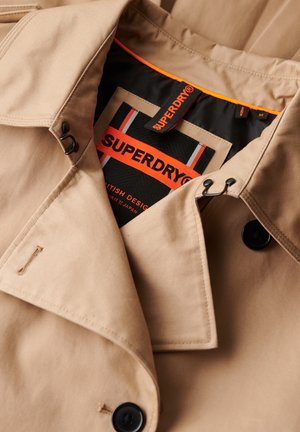 Gabán beige con botones oscuros, diseño de doble botonadura y forro de cuello negro. Presenta una etiqueta "SUPERDRY" en naranja y negro.