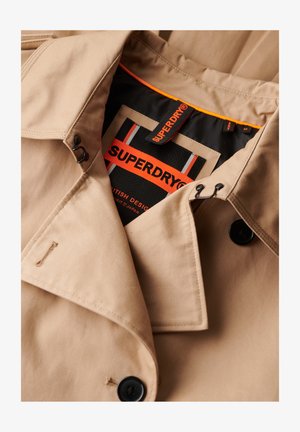 Beżowy płaszcz trench z ciemnymi guzikami, podwójnie zapięty design i czarną podszewką kołnierza. Posiada metkę "SUPERDRY" w pomarańczowym i czarnym kolorze.