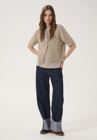 Beige strikket cardigan med korte ærmer og perleknapper, lagdelt over en stribet skjorte, parret med mørke denimbukser med frynser på ærmerne.