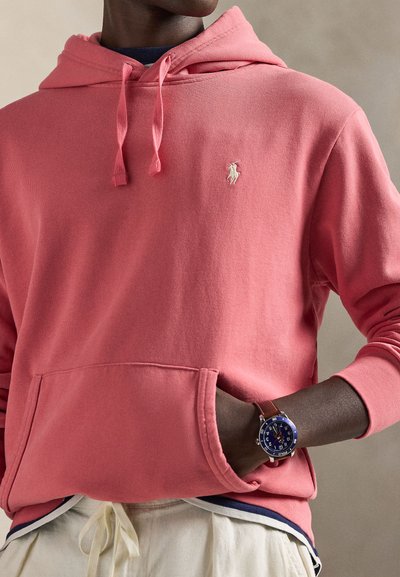 Κοραλί ροζ φούτερ με τσέπη μπροστά και κορδόνια, διακοσμημένο με ένα μικρό κεντητό λογότυπο. Καφέ ρολόι με μπλε καντράν στον καρπό.