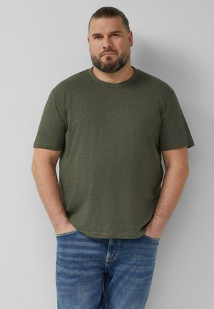 Mężczyzna z brodą i zaczesanymi do tyłu włosami stoi przy szarej ścianie, ubrany w oliwkowo-zieloną koszulkę i niebieskie dżinsy, ręce w kieszeniach.