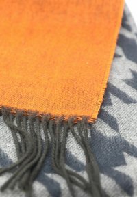 Geweven sjaal met een oranje deel met franjes en een contrasterende grijze achtergrond met een hounds-tooth patroon. Zachte textuur.