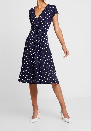 Marineblauwe wikkeljurk met witte polkadots, korte mouwen en een ceintuur in de taille. De vloeiende rok reikt tot halverwege de kuit en is gemaakt van lichtgewicht stof.