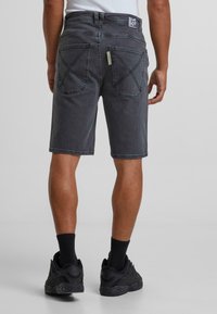 Shorts en denim noir avec une coupe droite, dotés de poches arrière à double couture et d'un accent en label jaune. Portés avec des baskets noires et des chaussettes.