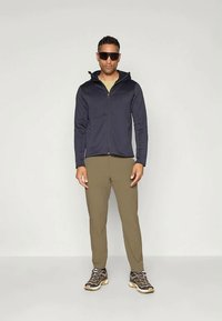 Mees, kes kannab musta tõmblukuga kapuutsiga jakki, oliivrohelisi pükse, matkajalatsid ja suuri musti päikeseprille, seisab sileda tausta ees.