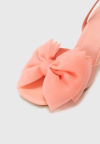 Rosa åpen tå slingback sandal med en stor, teksturert sløyfe laget av gjennomsiktig stoff. Skoen har en enkel, glatt såle og en lett hæl.