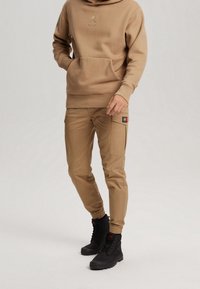 Persona con sudadera con capucha color beige y pantalones cargo con botas negras de cordones, de pie contra un fondo claro y liso.