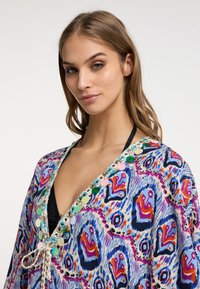 Šarena kaftan s vibrantnim apstraktnim uzorkom u plavoj, ružičastoj i narančastoj boji. Ukrašena je šljokicama i pletenicom sa vezanjem na izrezanju.