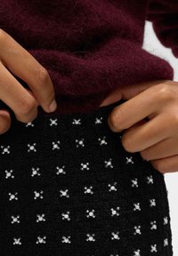 Schwarzer Stoffrock mit strukturierter Oberfläche, verziert mit silbernen Perlenakzenten in Blumenmustern. Dunkelburgunderfarbener Strickpullover ist teilweise sichtbar.