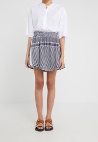 Camisa blanca de botones con manga corta, combinada con una falda azul marino con estampado geométrico. Se lleva con sandalias marrones.