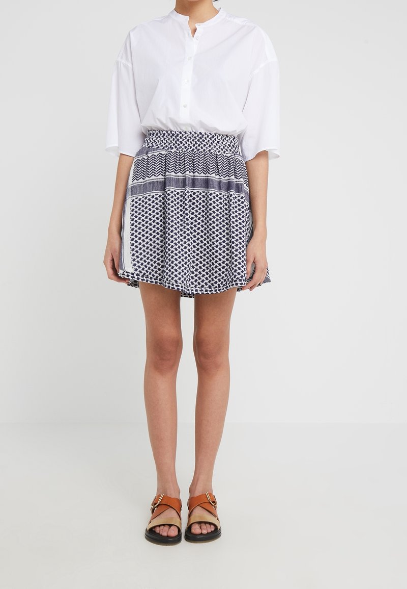Camisa blanca de botones con manga corta, combinada con una falda azul marino con estampado geométrico. Se lleva con sandalias marrones.