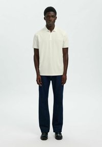 Jeune homme debout portant un polo blanc à manches courtes, un jean bleu foncé et des chaussures en cuir noir, sur un fond clair uni.