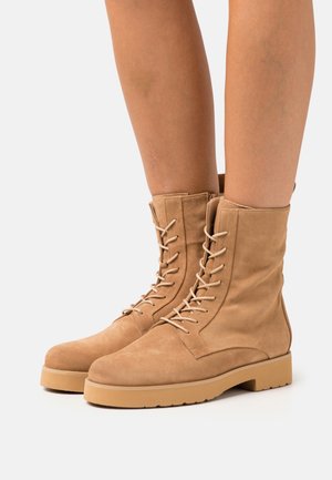 Botines de gamuza en tono beige con cordones, usados sobre piernas desnudas, con suelas planas texturizadas y cremalleras laterales, sobre un fondo blanco liso.