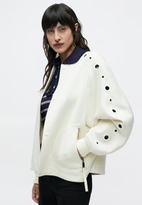 Γυναίκα με σκούρα μαλλιά, που φοράει λευκό σακάκι με πολλούς κυκλικούς ανοίγματα στο μανίκι, μπλε σκούρο πουκάμισο με γιακά και μαύρο παντελόνι.