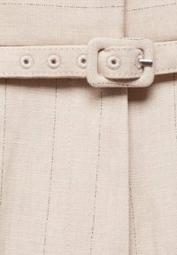 Gros plan sur une ceinture en tissu crème avec boucle rectangulaire et œillets cousus sur un vêtement assorti à texture similaire.