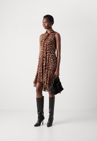Leopardprint mouwloos kleed met een strik-halslijn, uitlopende zoom en een doorschijnende afwerking. Gecombineerd met kniehoge zwarte leren laarzen en een zwarte tas.