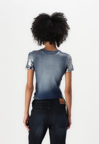 Shiny blue gradient short-sleeve top with a ribbed texture; worn with dark jeans featuring red accents and labels.