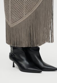 Μαύρες μυτερές μπότες με ψηλό τακούνι κάτω από φούστα σε χρώμα ταουπέ με κρόσσια, πλεκτή υφή και μακριές κρεμαστές κλωστές.