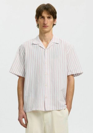 Ung mand iført en løs kortærmet skjorte med lodrette lyserøde og hvide striber samt lyse bukser foran en ensfarvet baggrund.